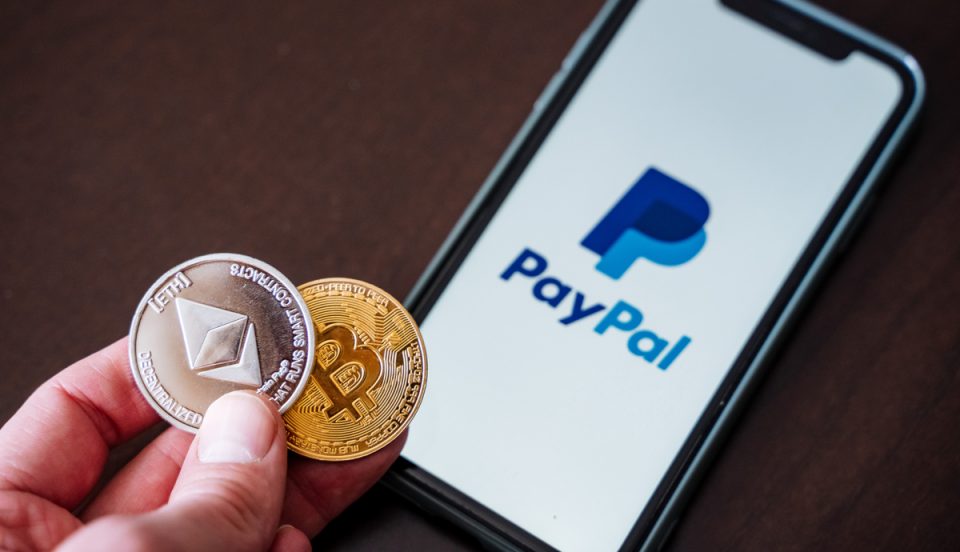 Mao segurando moedas de Ethereum e Bitcoin e um celular com o logo do PayPal ao fundo