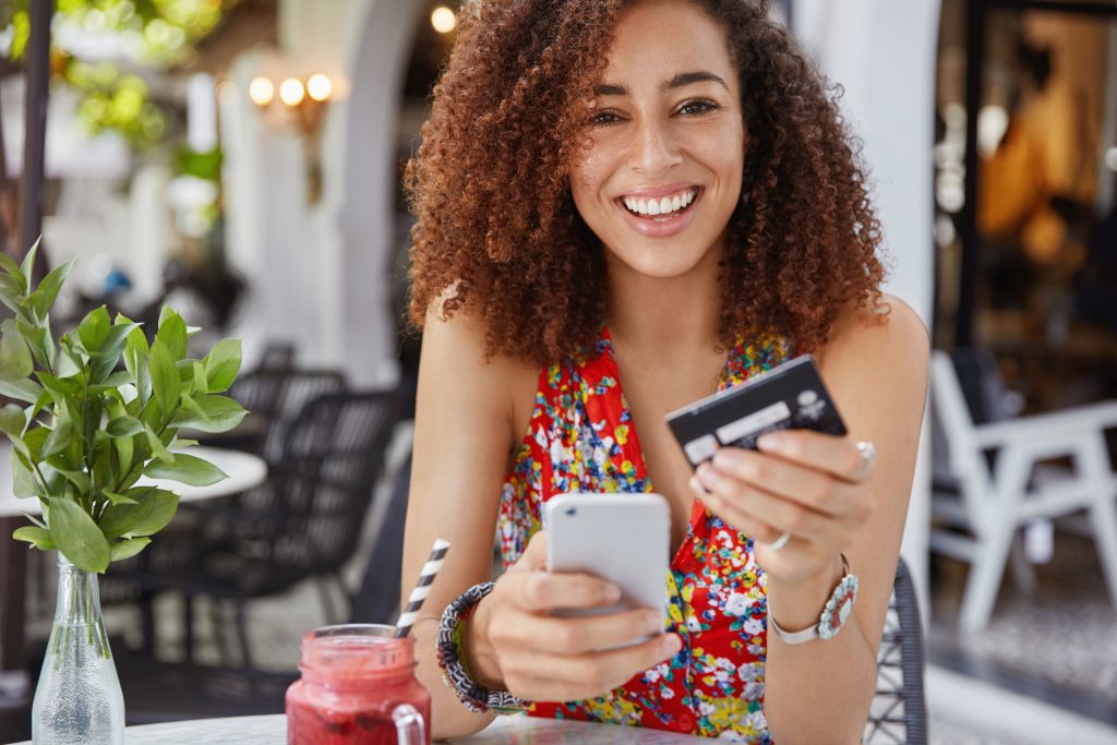 conceito de internet banking e comercio eletronico mulher jovem feliz e sorridente com penteado afro usa um celular moderno e um cartao de credito para fazer compras online 1
